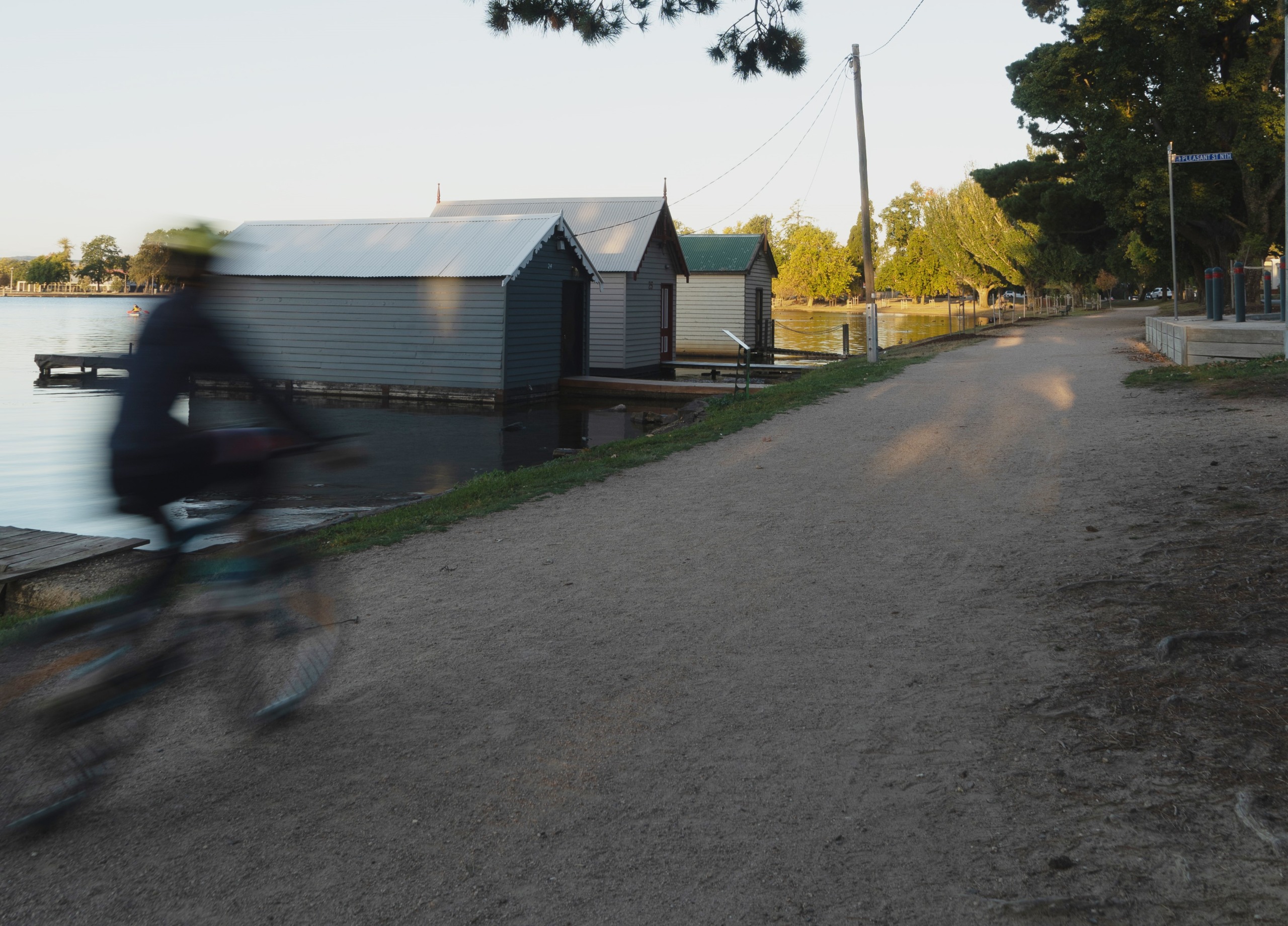 Cyclist riding past a row of boathouses along the southern shoreline of Lake Wendouree, with a gravel path and trees lining the foreshore.