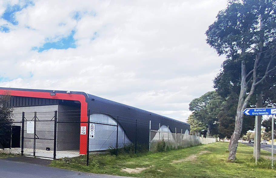 A modern building with a black and red exterior, surrounded by a fence, near a road sign pointing to "Ballarat Greyhounds."