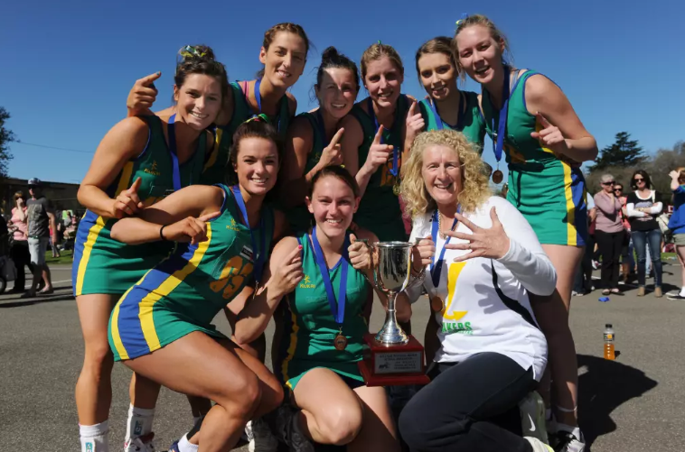 Sally McLean kneels with a Lake Wendouree netball team celebrating a premiership win. The players are wearing green and yellow uniforms, smiling and holding a trophy, with medals around their necks.