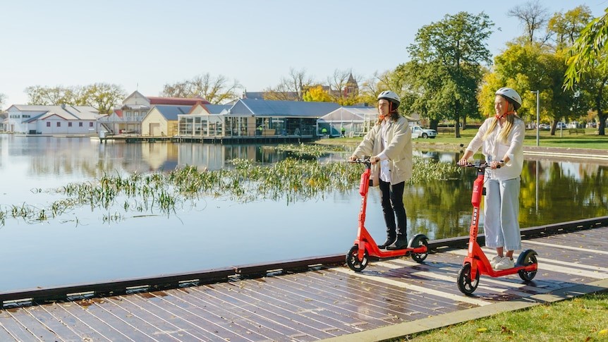 Two people riding bright orange shared e-scooters along the Lake Wendouree foreshore on a sunny day, wearing helmets and travelling on the wooden boardwalk beside the water.